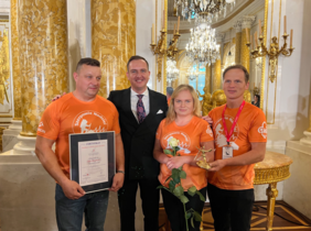 Pokaż zdjęcie: W Zamku Królewskim w warszawie do pamiątkowego zdjęcia pozują 4 osoby, 3 z nich ubrane są w podkoszulki w kolorze pomarańczowym z napisem Szerpowie Nadziei, pomiędzy nimi stoi mężczyzna w garniturze