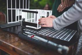 Pokaż zdjęcie: Na stole na pierwszym planie leży złożona laska dla osób niewidomych  obok stoi mini makieta scenografii przedstawienia. Widać fragment dłoni kobiety, która dotyka makietę.