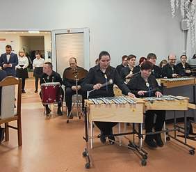 Pokaż zdjęcie: ystęp Na tym zdjęciu widać grupę muzyków, którzy występują podczas wydarzenia. Grają na różnych instrumentach, takich jak ksylofony i perkusja. Wszyscy są ubrani na czarno, co podkreśla formalny charakter występu. W tle znajdują się osoby obsługujące wydarzenie lub czekające na swoją kolej.