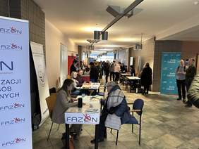 Pokaż zdjęcie: Za biurkiem siedzą dwie kobiety. Kobieta w ciemnych włosach czarnej koszuli, pracuje w laptopie. Druga w jaśniejszych niebieskiej koszuli i okularach trzyma w ręku dokumenty. Przed nimi siedzi mężczyzna w jeansach, bluzie i okularach. Za nimi kolejn