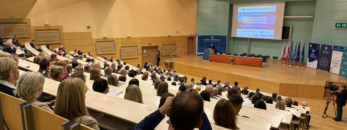 Pokaż zdjęcie: Sala wykładowa podczas konferencji. Uczestnicy siedzą na drewnianych ławkach w amfiteatralnym układzie. Na scenie prelegent przemawia na tle ekranu z prezentacją, a obok widoczne są flagi i bannery.