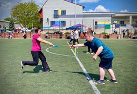 Pokaż zdjęcie: Na boisku wyłożonym sztuczną trawą znajduje się kobieta, która biegnąc przekazuje mężczyźnie niebieską pałeczkę. Na boisku znajdują się także inni zawodnicy, których dopingują kibice. W tle drzewa oraz budynek stanowiący kompleks sportowy. 