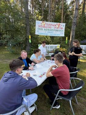 Pokaż zdjęcie: Grupa ośmiu osób znajduje się przy stoliku, na którym rozłożone zostały patyki do szaszłyków oraz kartki z grafikami prezentującymi różne owoce. W tle pomiędzy dwoma drzewami zawieszony jest baner z nazwą imprezy sportowo-integracyjnej.