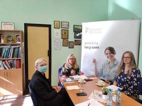 Pokaż zdjęcie: W pomieszczeniu przy dużym stole siedzą trzy kobiety, jedna z nich trzyma mikrofon. w tle stoi duży baner. Na białym tle jest logo PFRON, napis wszyscy jesteśmy tacy sami i młoda kobieta na wózku.