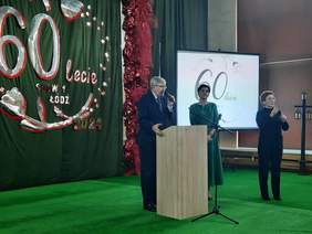 Pokaż zdjęcie: Na środku sali stoi przy mównicy mężczyzna, który przemawia do mikrofonu. Obok niego stoją dwie kobiety. Jedna z pań jest tłumaczem języka migowego. 