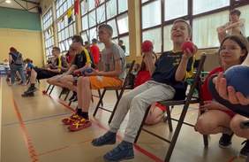 Pokaż zdjęcie: Na krzesłach w jednej linii siedzą dzieci trzymające piłki w rękach. Chłopiec na pierwszym kadrze uśmiecha się do aparatu. Obok niego kuca dziewczyna w okularach.