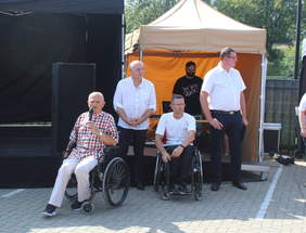 Pokaż zdjęcie: Zdjęcie przy scenie czterech mężczyzn w jasnych koszulkach, dwóch z nich siedzi na wózkach inwalidzkich, pozostałe osoby stoją z tyłu