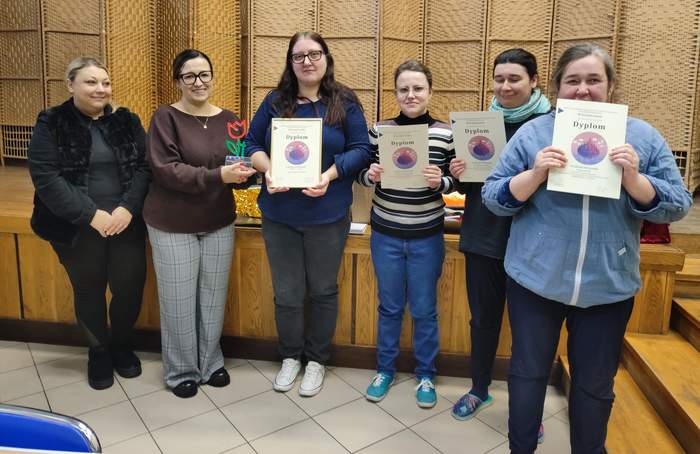 Pokaż zdjęcie: Grupa osób stojących na sali przed ażurową ścianką, trzymających dyplomy konkursowe. Jedna z osób wręcza symboliczny upominek w kształcie tulipana. Uczestnicy stoją w szeregu na tle podestu.