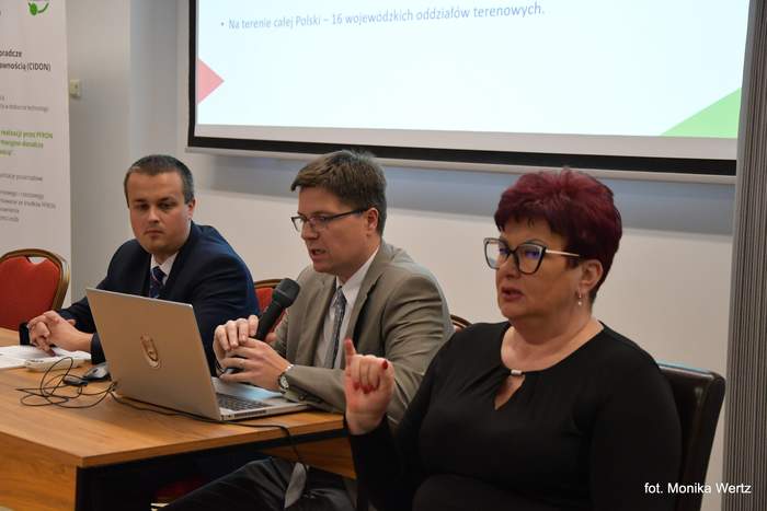 Pokaż zdjęcie: Dwóch pracowników Oddziału w trakcie konferencji, jeden z nich referuje i trzyma mikrofon, obok kobieta, który tłumaczy na język migowy