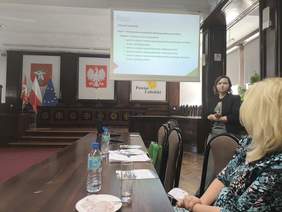 Pokaż zdjęcie: W sali konferencyjnej wykład prowadzi kobieta w ciemnej marynarce, w tle slajd z prezentacją, słuchacze siedzą przy stole ustawionym w podkowę.