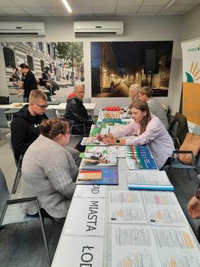 Pokaż zdjęcie: Przy stołach naprzeciwko siebie siedzą ludzie. Trwa Giełda Pracy dla Osób z Niepełnosprawnościami. Przy stoisku PFRON siedzi przedstawicielka, która rozmawia z kobietą i młodym mężczyzną. Na stole leżą ulotki promocyjne i materiały reklamowe PFRON. 