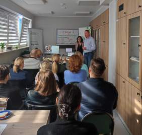 Pokaż zdjęcie: Grupa dorosłych uczestników siedzi w Sali  i słucha prelekcji prowadzonych przez kobietę i mężczyznę stojących przy tablicy. Na stoliku znajdują się dwa monitory wyświetlające slajdy prezentacji. Uczestnicy są uważni, niektórzy robią notatki. W pomieszczeniu jest jasne światło dzienne wpadające przez okna z żaluzjami. 