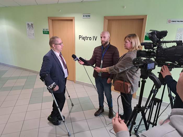 Pan Tomasz Wieczerzak, stoi o i udziela wywiadu, przed nim stoją reporterzy z mikrofonami, po prawej stronie widoczna kamera