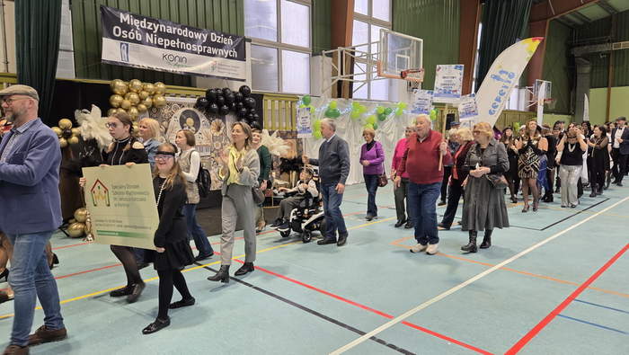 Pokaż zdjęcie: Zdjęcie przedstawia grupę ludzi poruszających się na hali sportowej  i trzymających transparenty. W tle widać dekoracje ze złotych, czarnych i zielonych balonów oraz baner wiszący na ścianie. Na banerze znajduje się napis: „Międzynarodowy Dzień Osób Niepełnosprawnych”.