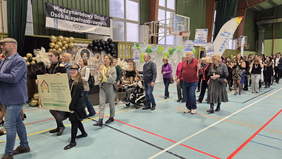 Pokaż zdjęcie: Zdjęcie przedstawia grupę ludzi poruszających się na hali sportowej  i trzymających transparenty. W tle widać dekoracje ze złotych, czarnych i zielonych balonów oraz baner wiszący na ścianie. Na banerze znajduje się napis: „Międzynarodowy Dzień Osób Niepełnosprawnych”.