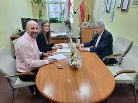 Pokaż zdjęcie: Przy owalnym stole siedzi kobieta i dwóch mężczyzn. Podpisują umowę. Na stole leżą różne dokumenty, długopisy, pieczątki, stroik świąteczny. 