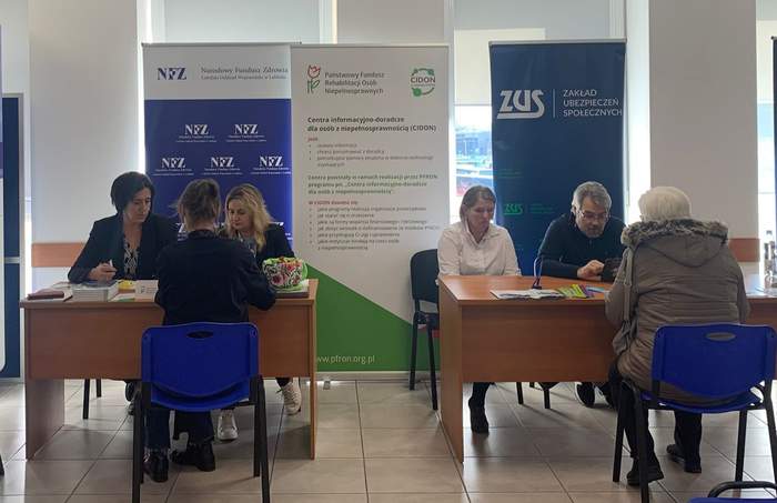 Pokaż zdjęcie: W sali przy dwóch biurkach siedzą  po dwie osoby z instytucji oraz osoby zainteresowane wsparciem. Na stolikach leżą foldery i ulotki. 