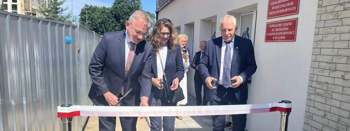 Pokaż zdjęcie: Przed białoczerwoną wstęgą stoi dwóch mężczyzn i kobieta pomiędzy nimi. Wszyscy ubrani na galowo trzymają w rękach nożyczki. Po prawej stronie nowy budynek z czerwonymi tablicami informacyjnymi. 