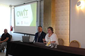 Pokaż zdjęcie: W sali konferencyjnej za stołem prezydialnym siedzi mężczyzna i kobieta, kobieta trzyma w ręku mikrofon, z lewej strony duży ekran z wyświetlaną prezentacjąą