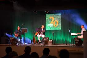 Pokaż zdjęcie: Na scenie znajdują się trzej mężczyźni, z czego dwóch gra na gitarze, a trzeci na pianinie. Jeden z nich śpiewa także do mikrofonu. W tle, na dużym ekranie wyświetlone jest sformułowanie: „20-lecie Środowiskowego Domu Samopomocy w Ślesinie”.