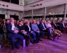 Pokaż zdjęcie: Grupa kilkudziesięciu osób siedzi w dużej sali i jest oświetlona światłami reflektorów. Część z tych osób ma ze sobą różne upominki, w tym między innymi kwiaty doniczkowe.