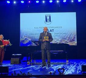 Pokaż zdjęcie: Ujęcie ze środka widowni, przedstawiające mężczyznę w ciemnym garniturze stojącego na oświetlonej na niebiesko scenie. Mężczyzna trzyma złotą statuetkę w kształcie serca i stoi przed mikrofonem. Po jego lewej stronie znajduje się kobieta z bukietem kwiatów. W tle widać ekran z tym samym napisem, co na zdjęciu (3).jpg, dotyczącym 25 lat ZAZ "CENTRALNA KUCHNIA" Koła PSONI w Stargardzie. Na pierwszym planie widoczne są fragmenty głów osób siedzących najbliżej.