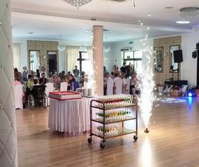 Pokaż zdjęcie: Sala bankietowa, w centrum sali stoi stolik z tortem i fajerwerkami. W tle uroczyście zastawione stoły, przy których siedzi kilka osób.