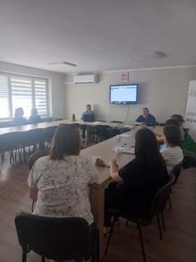 Pokaż zdjęcie: Kilka osób siedzi przy stole. Przed nimi wyświetlana jest prezentacja. Po prawej stronie baner PFRON.