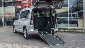 Pokaż zdjęcie: Auto dostosowane do potrzeb osoby z niepełnosprawnością na wózku inwalidzkim, otwarty tył auta, z windą wjazdową, auto stoi na tle salonu samochodowego