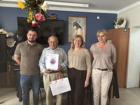 Pokaż zdjęcie: Cztery osoby stoją obok siebie w przytulnym pomieszczeniu przypominającym salon lub pokój dzienny. Jedna z osób trzyma dyplom oraz torbę z upominkami. W tle widoczne są ciemna ściana, telewizor, obraz oraz drewniane meble. Uczestnicy stoją blisko siebie i uśmiechają się do zdjęcia wykonanego podczas uroczystego wręczenia wyróżnienia.
