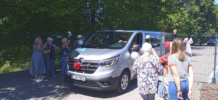 Pokaż zdjęcie: Srebrny bus do przewozu osób z niepełnosprawnościami, a obok podopieczni OPS, jest słonecznie