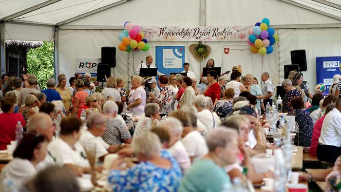 Pokaż zdjęcie: Zdjęcie grupy osób siedzącej pod dużym namiotem, scena przyozdobiona jest balonami, goście patrzą w kierunku sceny, na której trwa występ zespołu muzycznego - cztery osoby.