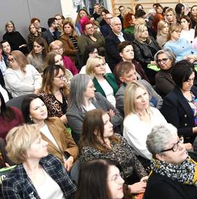 Pokaż zdjęcie: Zbliżenie na osoby będące uczestnikami konferencji, siedzące na krzesełkach w sali gimnastycznej.