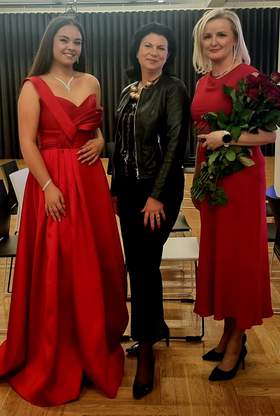 Trzy kobiety na uroczystej Gali, dwie ubrane w długie czerwone suknie, jedna ubrana na czarno pozują do zdjęcia.