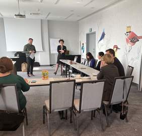 Pokaż zdjęcie: w sali szkoleniowej przy stołach siedzi kilka osób robią notatki ze spotkania, na przeciw ich stoi mężczyzna i kobieta.