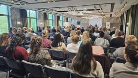 Pokaż zdjęcie: Grupa kilkudziesięciu osób znajduje się na sali konferencyjnej i przysłuchuje się wystąpieniu dwóch mężczyzn. Na dużym ekranie wyświetlona jest prezentacja, a spotkanie tłumaczone jest na język migowy.