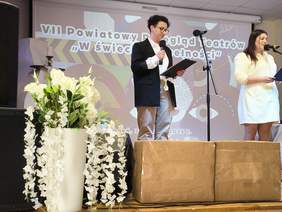 Pokaż zdjęcie: Na scenie wydarzenia dwie osoby stoją przy mikrofonach i czytają z kart. W tle ekran z napisem „VII Powiatowy Przegląd Teatrów »W świecie niepełnosprawności«”. Obok widoczna dekoracja kwiatowa i elementy scenografii.