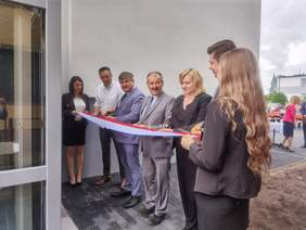 Pokaż zdjęcie: kilka osób stoi na zewnątrz trzyma szarfę biało-czerwoną jedna z osób przecina ją nożyczkami 