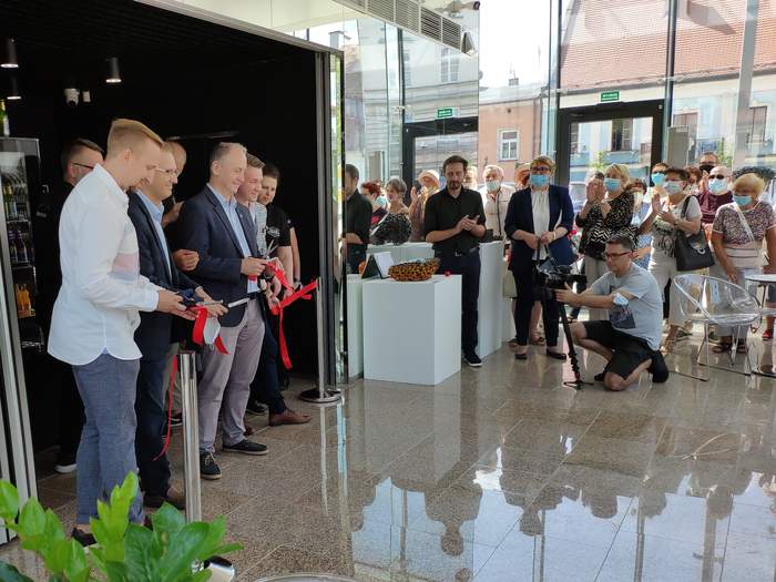 Pokaż zdjęcie: Wewnątrz kawiarni w szeregu stoi kilku mężczyzn, trzymają w rękach kawałki czerwonej wstęgi którą przecięli, obserwuje ich grupa ludzi