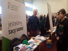 Pokaż zdjęcie: na zdjęciu w tle baner z logo PFRON, obok stoi mężczyzna i przekazuje informacje, na stoliku są umieszczone materiały i ulotki o dofinansowaniach ze środków PFRON, dookoła tłum młodych ludzi