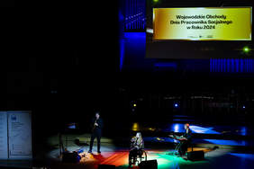 Pokaż zdjęcie: Na wózku inwalidzkim siedzi kobieta, która śpiewa do mikrofonu. Znajduje się na scenie Filharmonii Łódzkiej. Za kobietą stoją dwie osoby, które grają na instrumentach muzycznych.