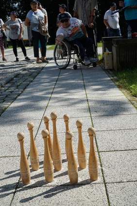 Pokaż zdjęcie: Młody chłopak na wózku inwalidzkim celuje w drewniane kręgle ustawione na chodniku 