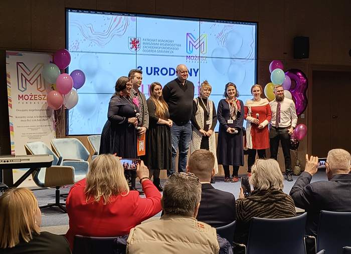 Pokaż zdjęcie: Grupa osób stojąca na scenie w sali Delfin w siedzibie Urzędu Marszałkowskiego Województwa Zachodniopomorskiego podczas uroczystości trzecich urodzin Fundacji MOŻESZ. W tle widać logo fundacji, balony oraz ekran z napisem „3 URODZINY”. Publiczność siedzi na krzesłach, robiąc zdjęcia i obserwując wydarzenie.