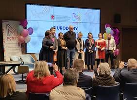 Pokaż zdjęcie: Grupa osób stojąca na scenie w sali Delfin w siedzibie Urzędu Marszałkowskiego Województwa Zachodniopomorskiego podczas uroczystości trzecich urodzin Fundacji MOŻESZ. W tle widać logo fundacji, balony oraz ekran z napisem „3 URODZINY”. Publiczność siedzi na krzesłach, robiąc zdjęcia i obserwując wydarzenie.