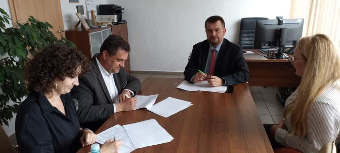 Pokaż zdjęcie: Dwóch mężczyzn i dwie kobiety siedzą przy biurku w trakcie podpisywania umów w pomieszczeniu Oddziału, wszyscy są ubrani na galowo