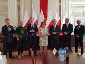 siedem osób, dwie kobiety i pięciu mężczyzn, elegancko ubrani stoja w szeregu na tle biało-czerwonych flag, mężczyzna z lewej przemawia