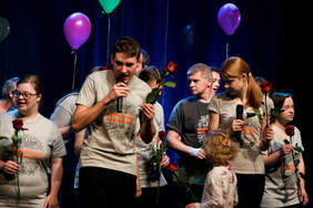 Pokaż zdjęcie: Zdjęcie sceny, na scenie około dwunastu osób. Są to kobiety, dzieci i mężczyźni. Na pierwszym planie mężczyzna z mikrofonem śpiewa, kobieta trzymają jedną różę w ręku.