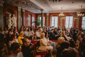 Pokaż zdjęcie: Sala konferencyjna z publicznością siedzącą na krzesłach twarzą do sceny. Wokół sali stoją wysokie kolumny. Po lewej stronie widoczna jest duża, jasna rzeźba dwóch postaci ludzkich na postumencie. Ściany mają ciepły, czerwony kolor.