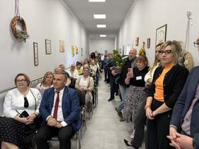 Pokaż zdjęcie: Grupa osób siedzących i stojących w długim korytarzu podczas wydarzenia. W tle dekoracje ścienne. Atmosfera jest oficjalna.