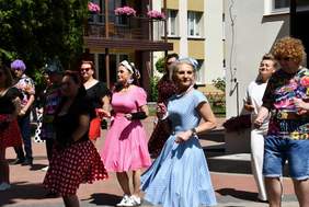 Pokaż zdjęcie: Grupa jedenastu osób, zarówno kobiet, jak i mężczyzn ubrana jest w barwne stroje i porusza się krokiem tanecznym będąc zwrócona w jedną stronę. W tle zabudowania, drzewa, krzewy i kwiaty.